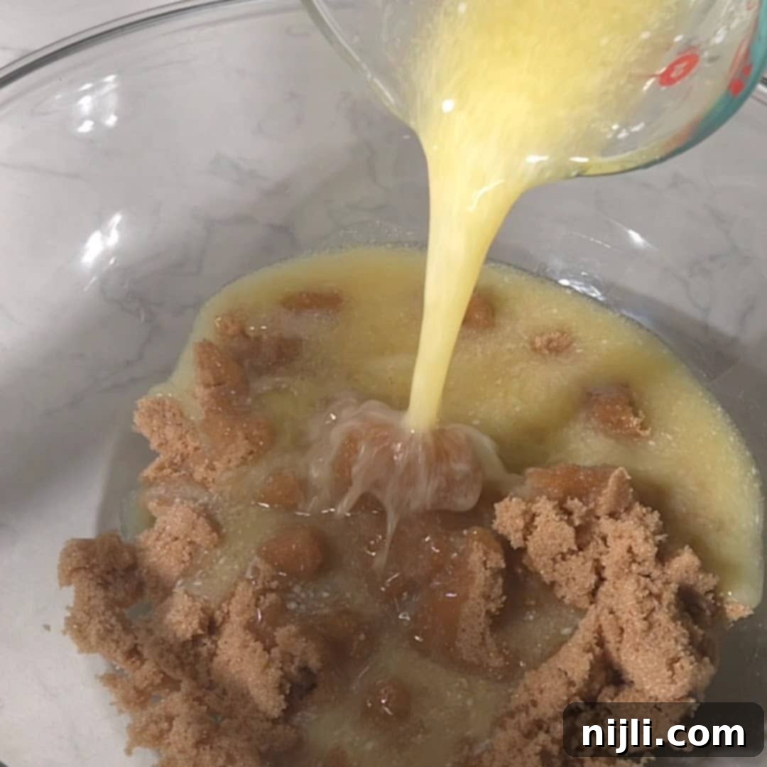 M&M Burst Chocolate Chip Goodness 8 A bowl with brown sugar as melted butter is poured over it, beginning the process of mixing for chocolate chip M&M cookies. This step softens the sugar and butter mixture, creating a rich, creamy base essential for the cookies' chewy texture.