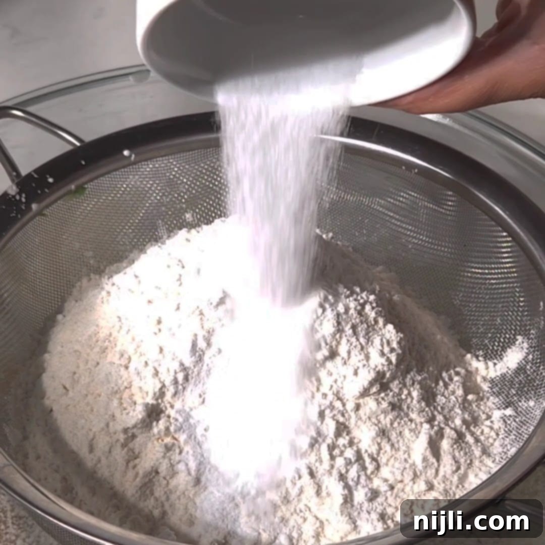 M&M Burst Chocolate Chip Goodness 10 White flour being sifted through a fine mesh strainer into a mixing bowl, helping to remove any clumps and aerate the flour for a smoother M&M cookie dough texture. Sifting the flour is a crucial step in creating soft and fluffy cookies.