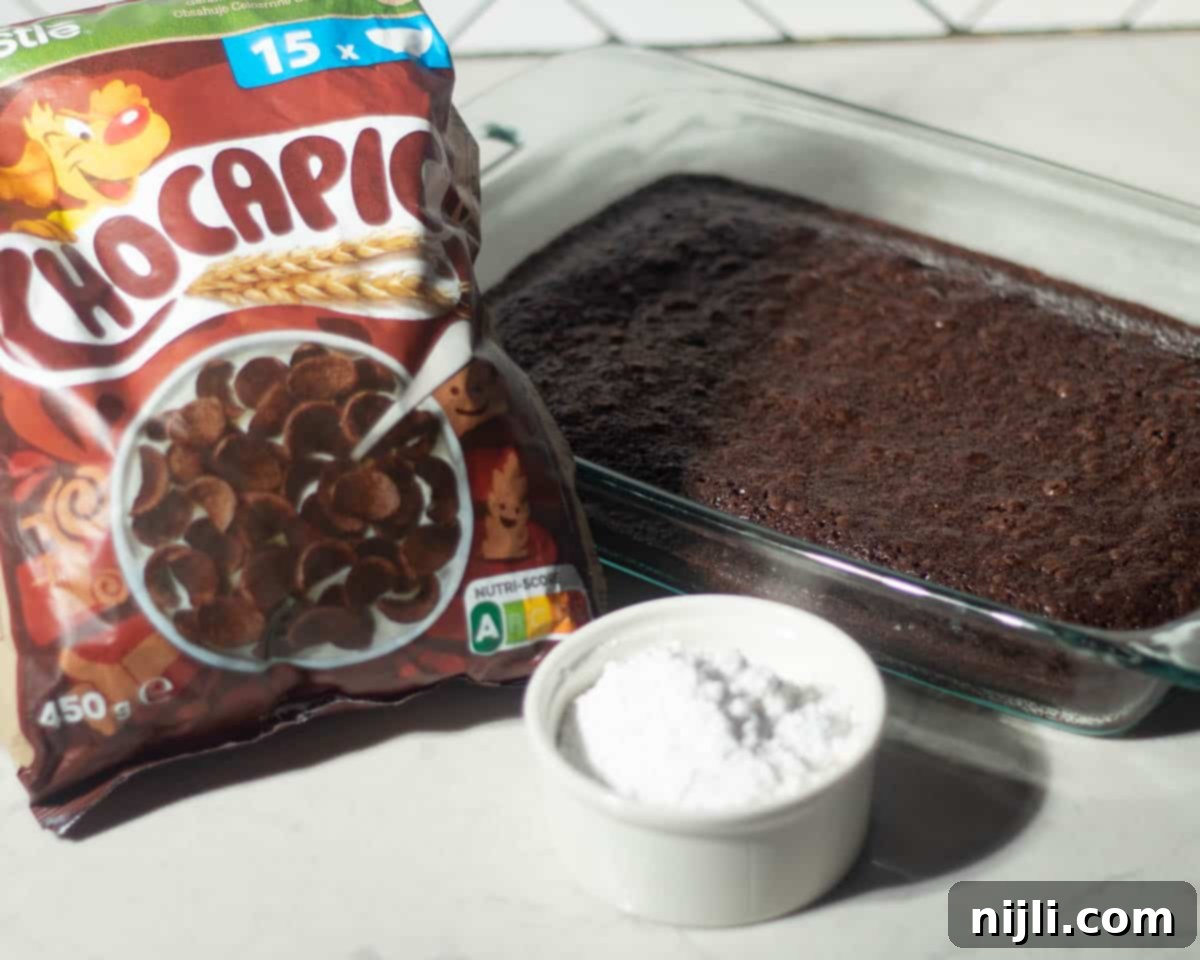 Delicious Pinecone Brownie Recipe 4 A flat-lay shot of ingredients for pinecone brownies, including a baked brownie slab, powdered sugar in a ramekin, and a bag of chocolate cereal.