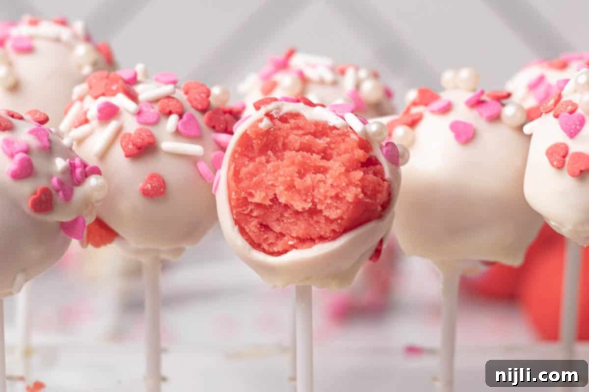 Sweetheart Cake Pops 12 A display of Valentine's cake pops coated in white chocolate and decorated with pink and red heart-shaped sprinkles. The moist pink cake inside of one pop is revealed, offering inspiration for festive Valentine's Day treats.