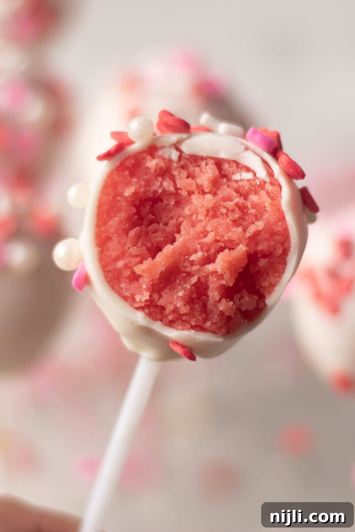 Sweetheart Cake Pops 13 A single Valentine's cake pop held by hand, featuring a smooth white chocolate coating with pink and red heart-shaped sprinkles. A bite reveals its soft pink cake center, making it a perfect dessert for Valentine's Day.