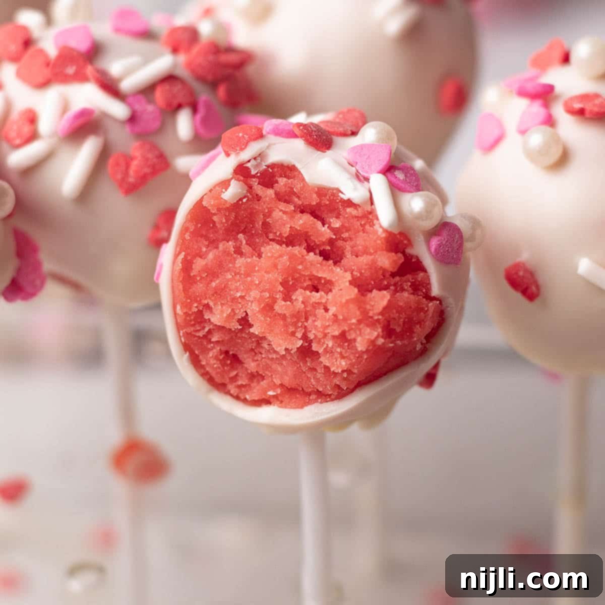 Sweetheart Cake Pops 15 Valentine's Day cake pops on display, with one cut open to reveal its moist pink interior. The white chocolate coating is decorated with red and pink heart-shaped sprinkles, adding a festive touch.