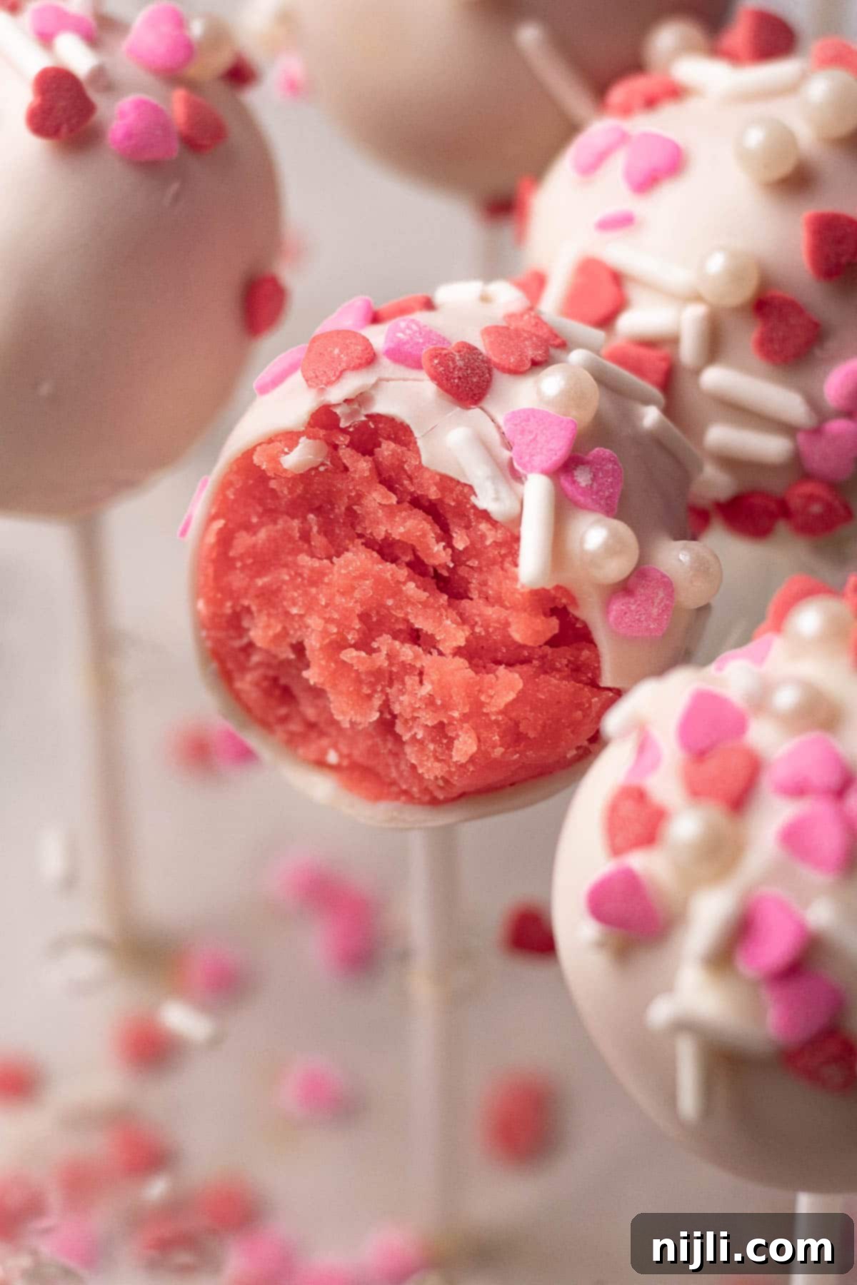 Sweetheart Cake Pops 3 Valentine's cake pops on display, coated in white chocolate and sprinkled with pink and red hearts. One pop is cut open to reveal its moist pink cake interior, highlighting a sweet Valentine's Day dessert idea.
