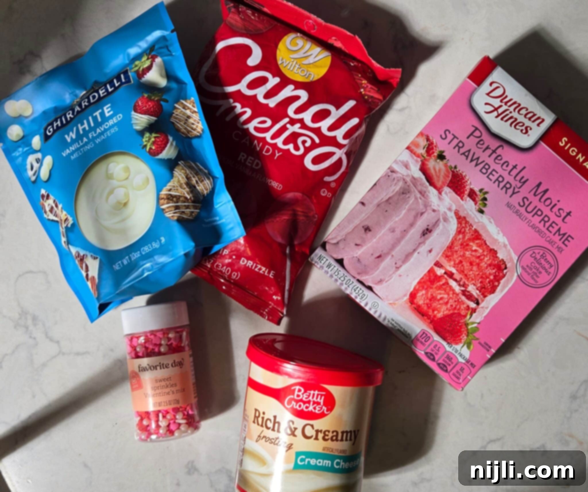 Sweetheart Cake Pops 4 A flat lay of ingredients for making Valentine's cake pops, including Ghirardelli white melting wafers, Wilton red candy melts, Duncan Hines Strawberry Supreme cake mix, Betty Crocker cream cheese frosting, and a jar of Valentine's-themed sprinkles. Perfect for creating easy and festive Valentine's Day desserts.