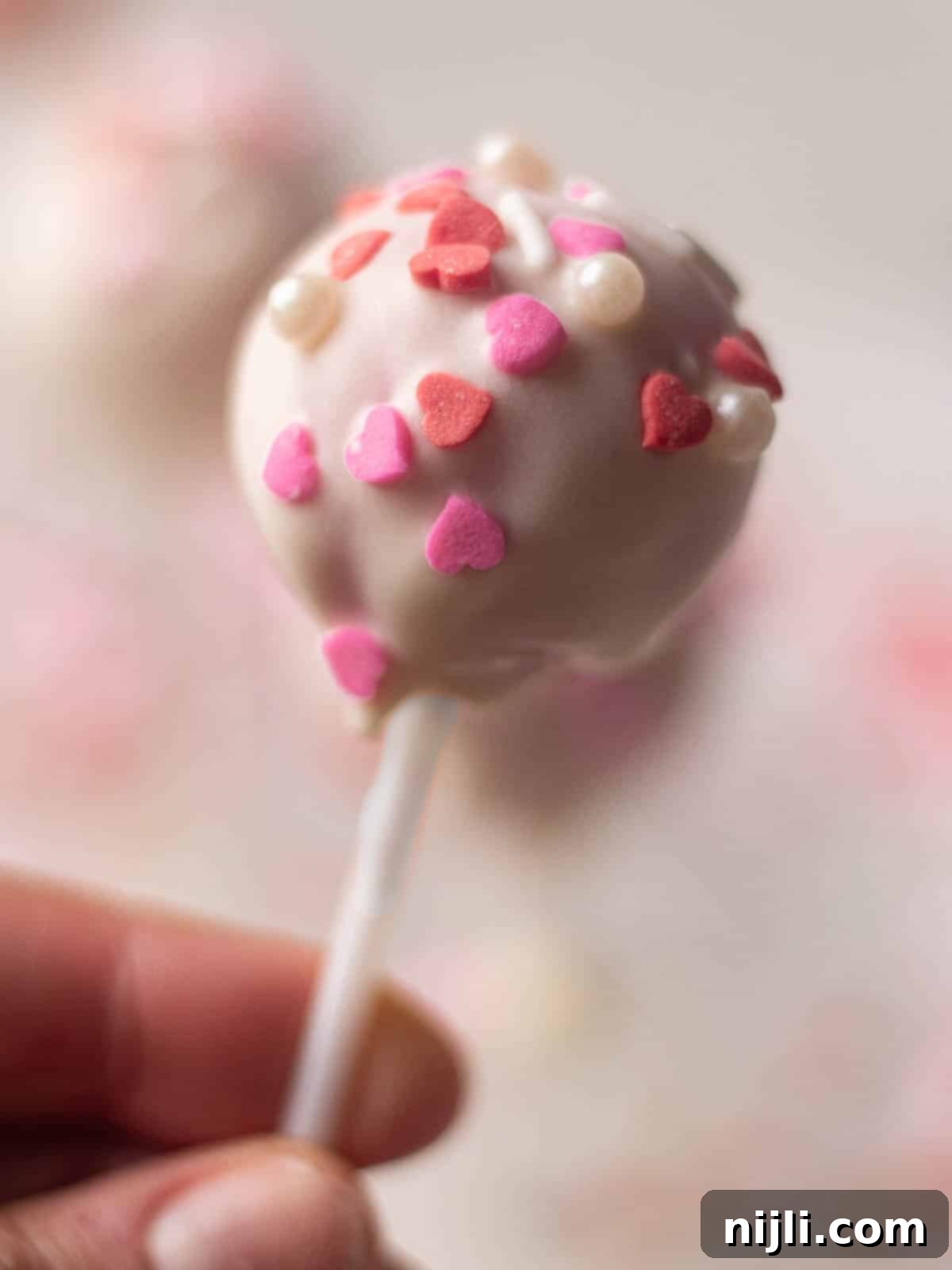 Sweetheart Cake Pops 5 A close-up of a Valentine's cake pop with white chocolate coating and pink and red heart-shaped sprinkles. A bite taken out of the pop reveals the vibrant pink cake inside, showcasing a delicious and festive treat idea for Valentine's Day.