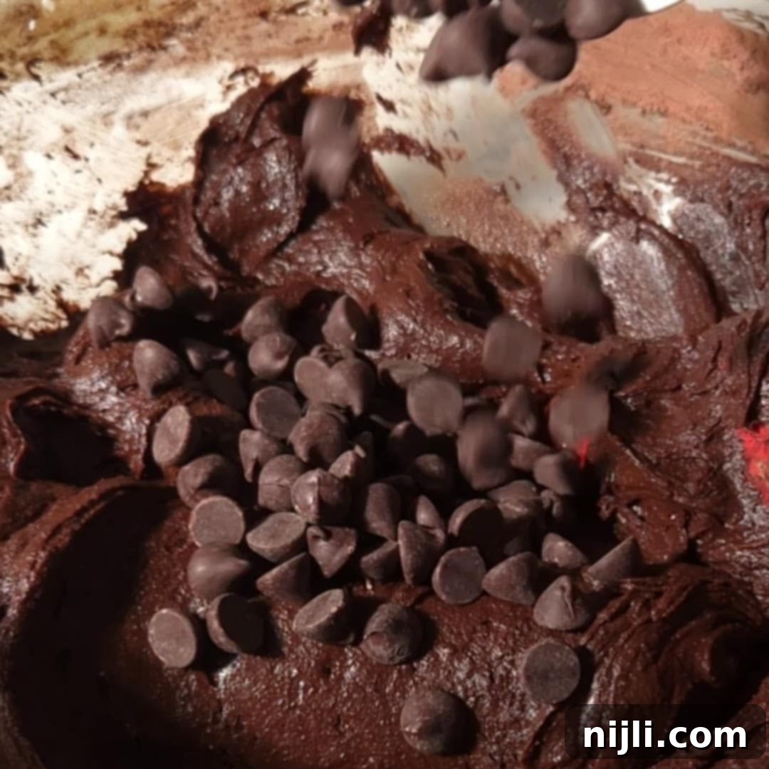 Chocolate chips being folded into rich brownie batter, creating a chunky texture perfect for fudgy brownies with cream cheese frosting. The focus is on the indulgent batter ready for baking.