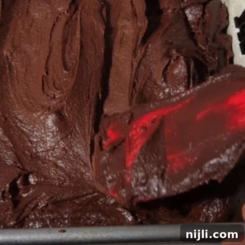 A close-up of brownie batter being spread evenly in a baking pan with a red spatula, preparing the base for brownies with cream cheese frosting. The smooth batter hints at a decadent and chocolatey treat.