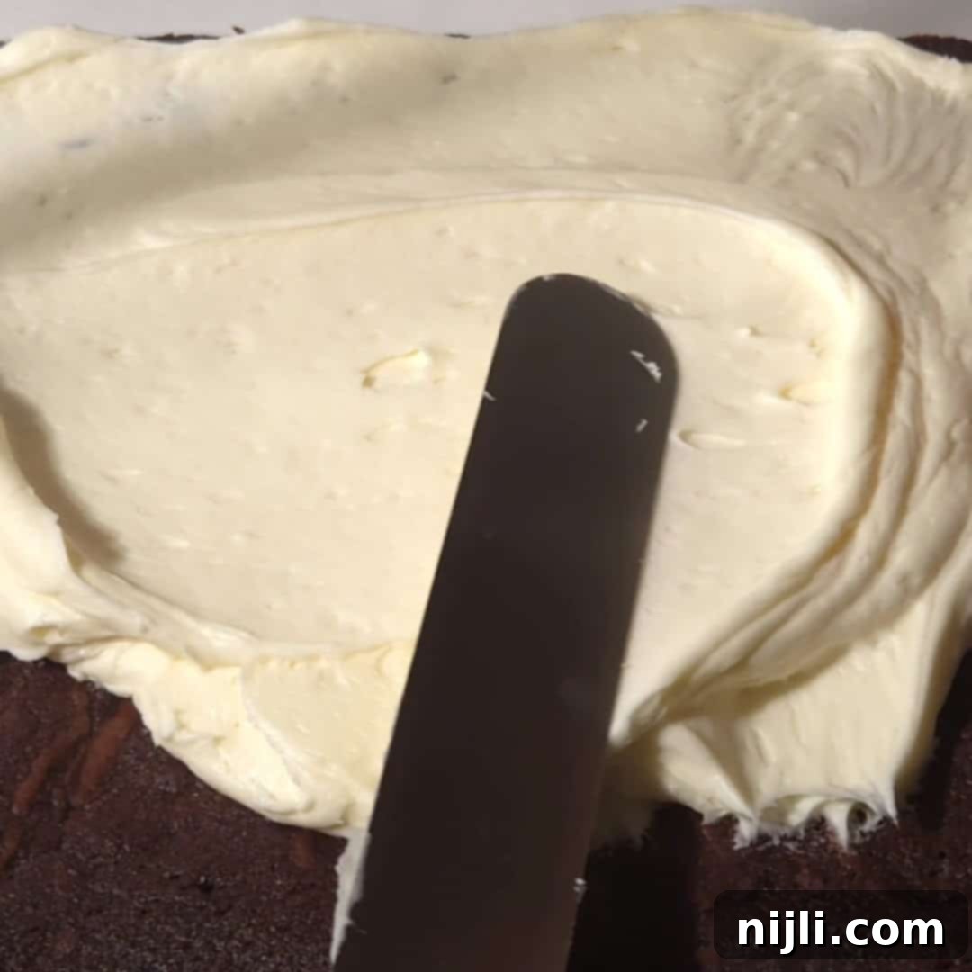 A close-up of a spatula spreading smooth cream cheese frosting on freshly baked chocolate brownies, showcasing the rich, velvety texture of the topping.