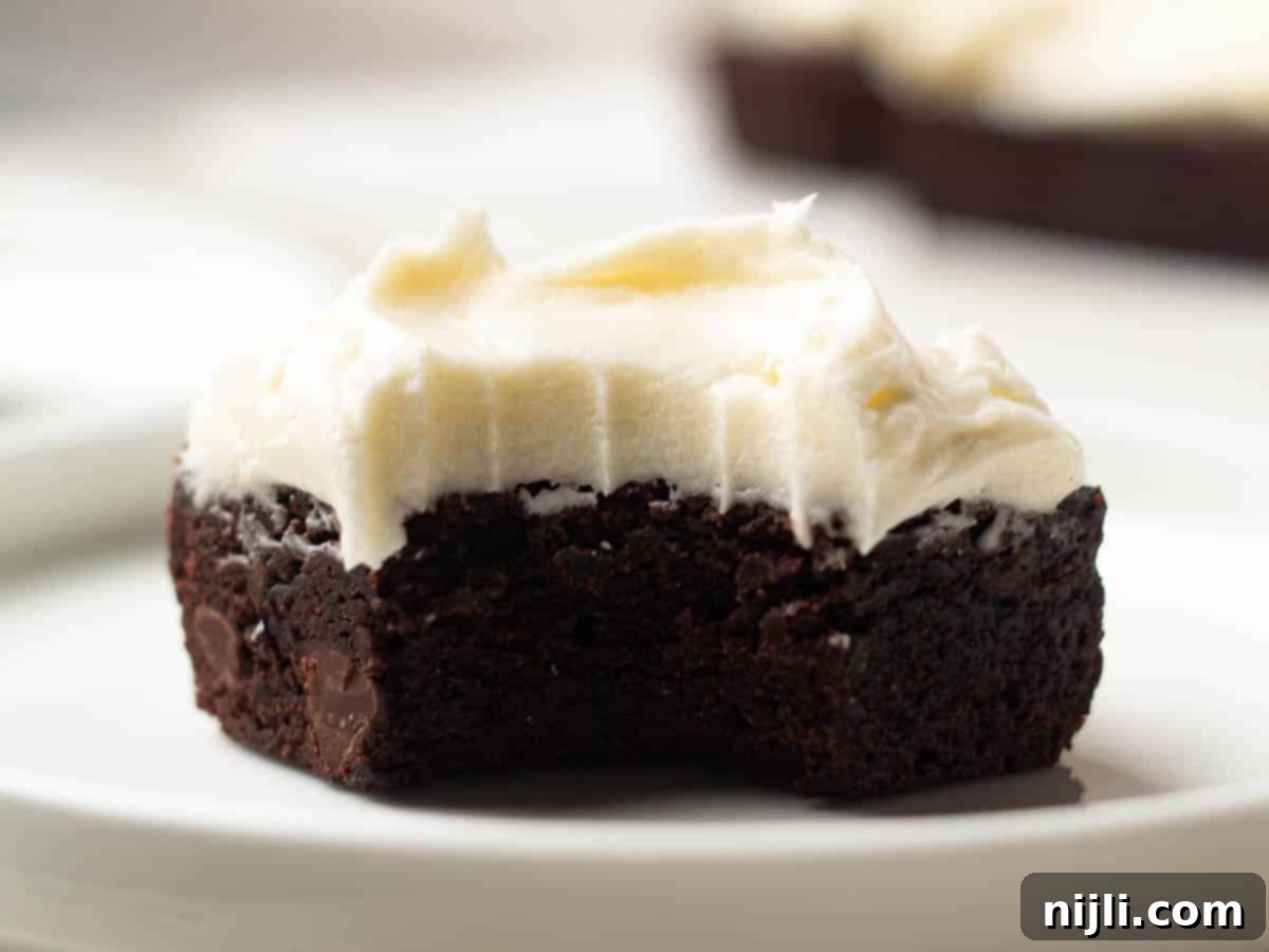 A partially eaten brownie topped with cream cheese frosting, revealing its moist, chocolatey center.
