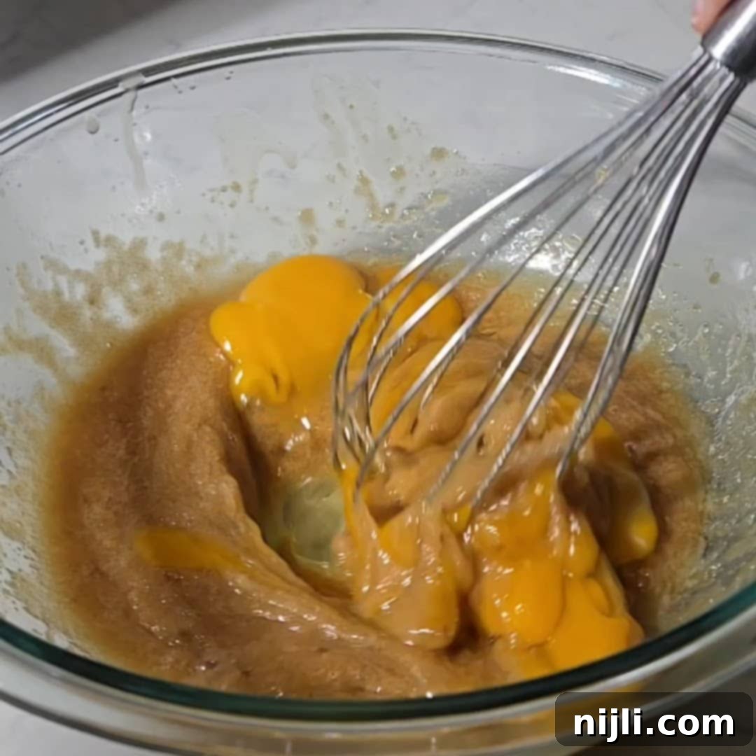 A whisk blending eggs into sugar and butter, creating a smooth mixture for brownie batter. This step builds the rich base for brownies with cream cheese frosting.