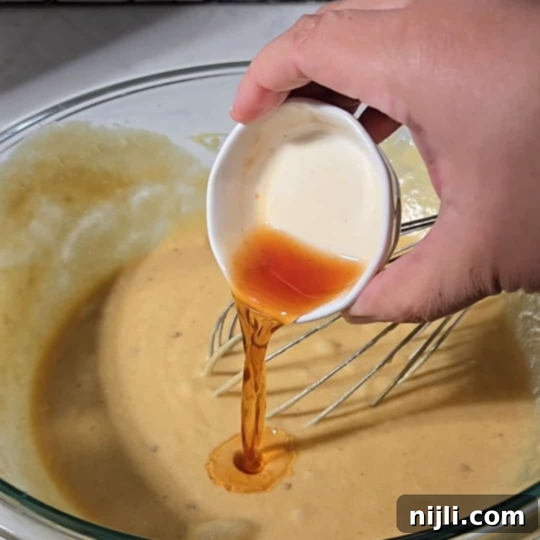 A small bowl of vanilla extract being poured into wet ingredients, combining to create the flavorful base for brownies with cream cheese frosting. The golden liquid adds a warm, aromatic touch.