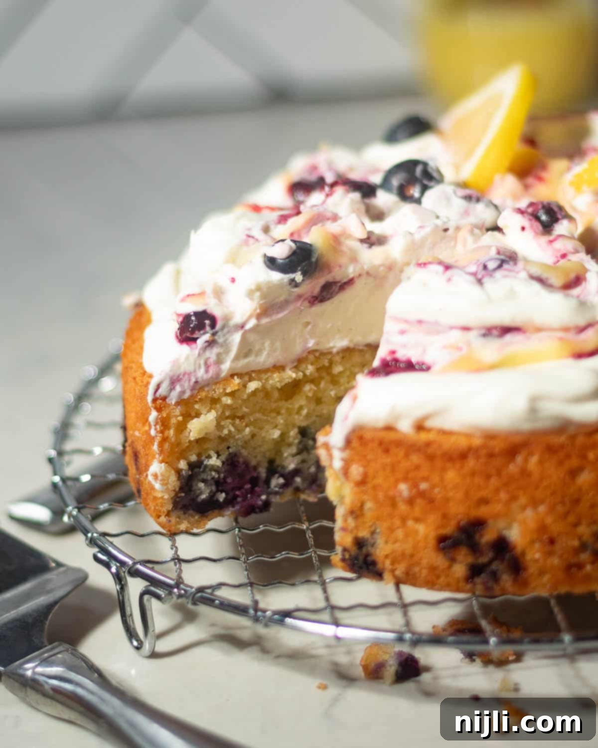 Sunny Lemon Blueberry Mascarpone Delight 3 Side view of a sliced lemon blueberry mascarpone cake on a wire rack, showcasing the cake’s golden crumb, embedded blueberries, and rich mascarpone topping.