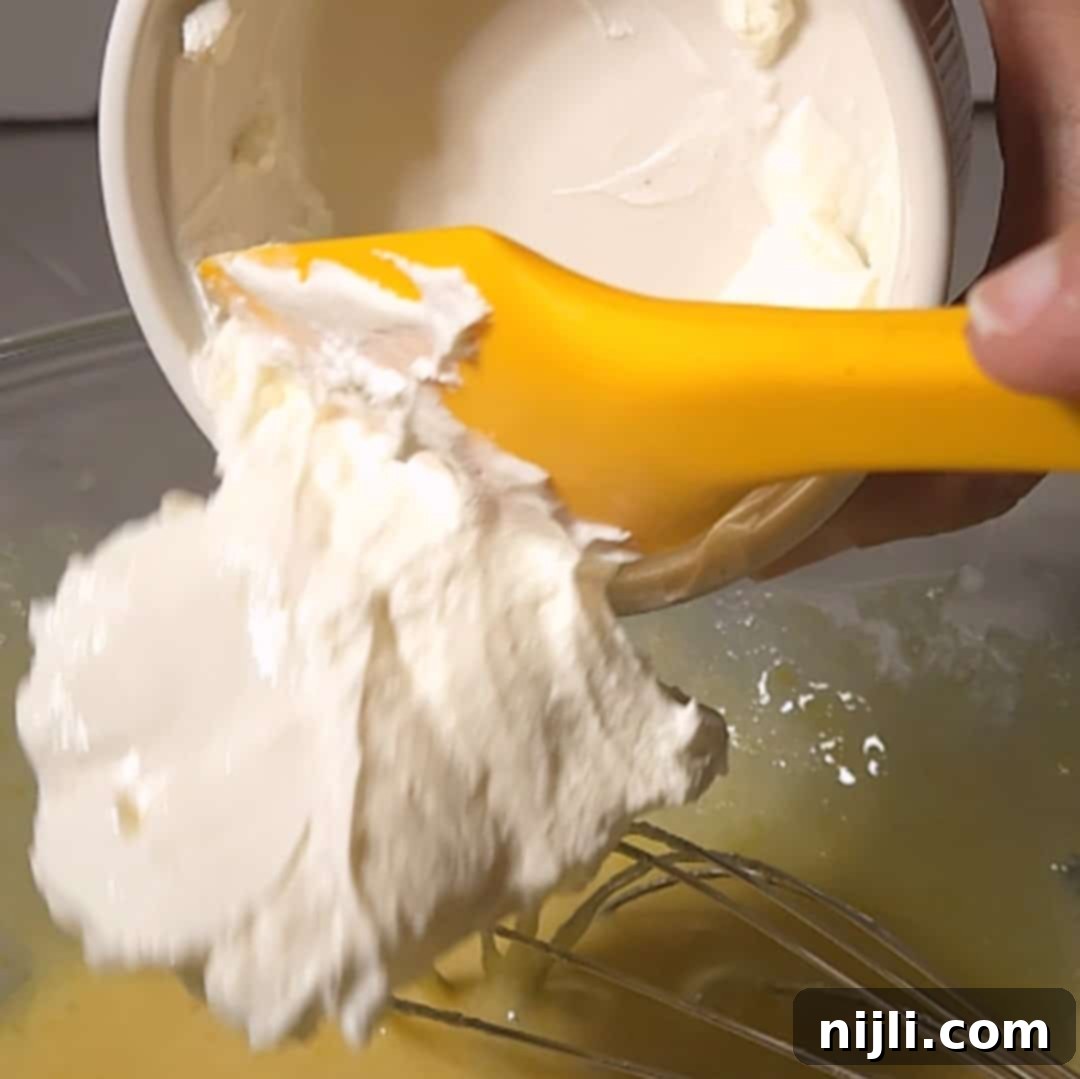 Sunny Lemon Blueberry Mascarpone Delight 9 A yellow spatula scooping mascarpone cheese from a ramekin into a bowl of mixed wet ingredients, showing a key step in making a lemon blueberry mascarpone cake batter.