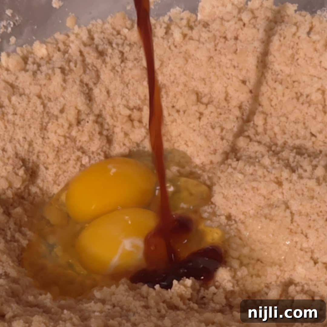 Homemade Blackberry Coffee Cake 10 Two eggs and vanilla extract are added to a bowl of crumbly flour mix, forming the wet ingredients for blackberry coffee cake batter.