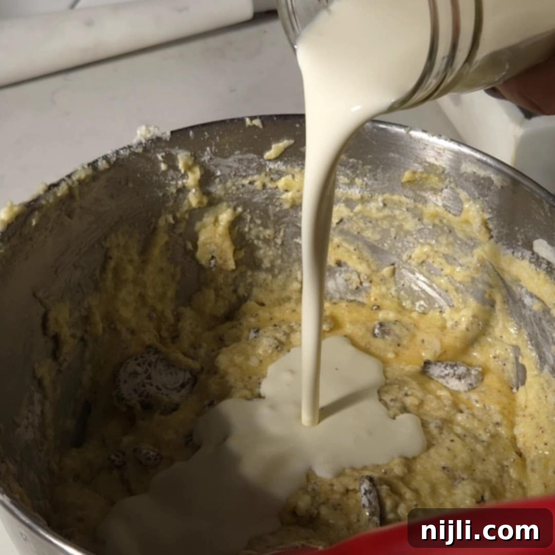 Decadent Oreo Pound Cake 11 Buttermilk being poured into the Oreo pound cake batter in a mixing bowl, partially blended with visible chunks of Oreo cookies.