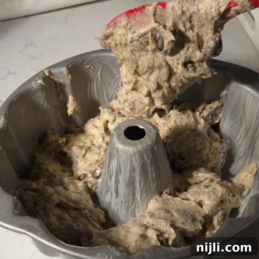 Decadent Oreo Pound Cake 12 Thick Oreo pound cake batter being spooned into a greased Bundt pan, filled with chunks of Oreo cookies.