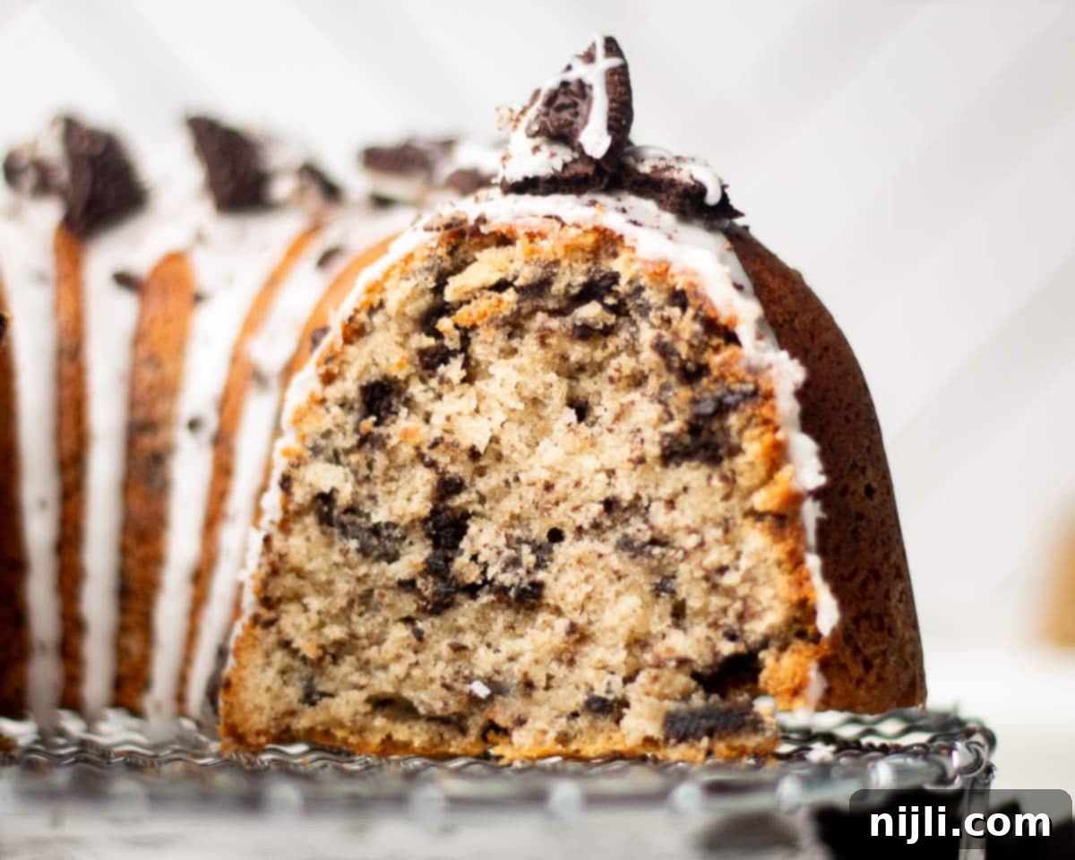 Decadent Oreo Pound Cake 3 Front view of a thick Oreo pound cake slice topped with glaze and crushed Oreos, with the rest of the sliced Bundt cake blurred in the background.