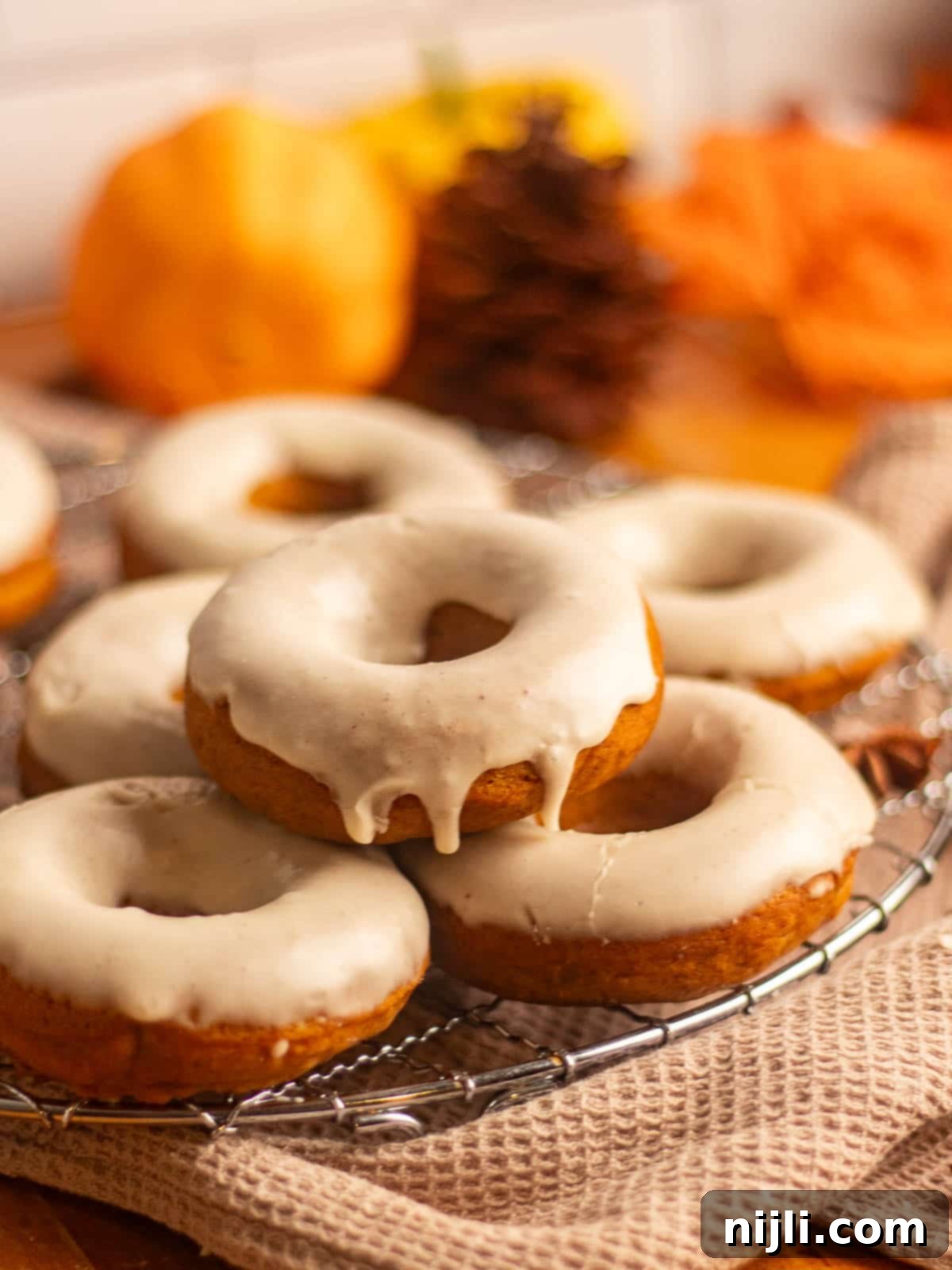 Spiced Pumpkin Perfection 7 Pumpkin spice donuts stacked on a cooling rack, with glaze glistening under soft lighting and pumpkins blurred in the background.