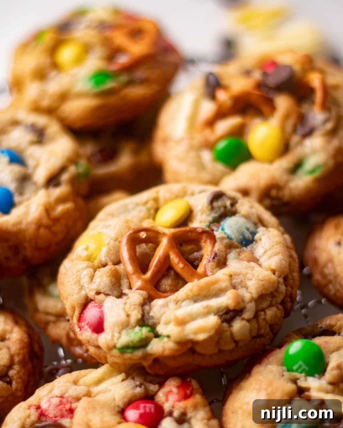 The Everything Cookie 3 Cooling rack of chewy homemade kitchen sink cookies with pretzels, potato chips, and chocolate candies scattered around.