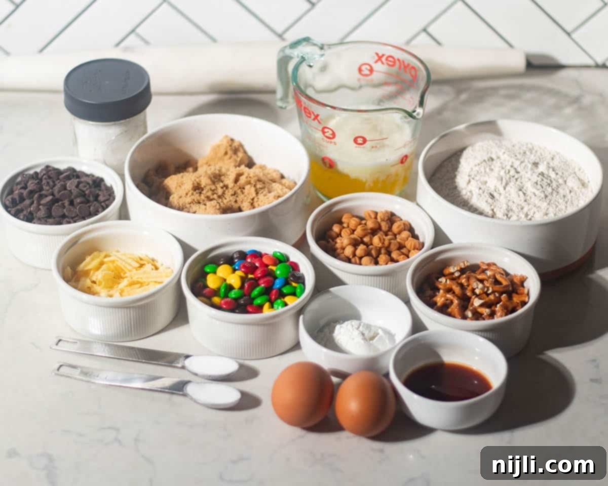 The Everything Cookie 5 Overhead shot of kitchen sink cookie ingredients including flour, brown sugar, melted butter, chocolate chips, M&Ms, pretzels, potato chips, peanut butter chips, eggs, vanilla, baking soda, and salt arranged on a marble counter.