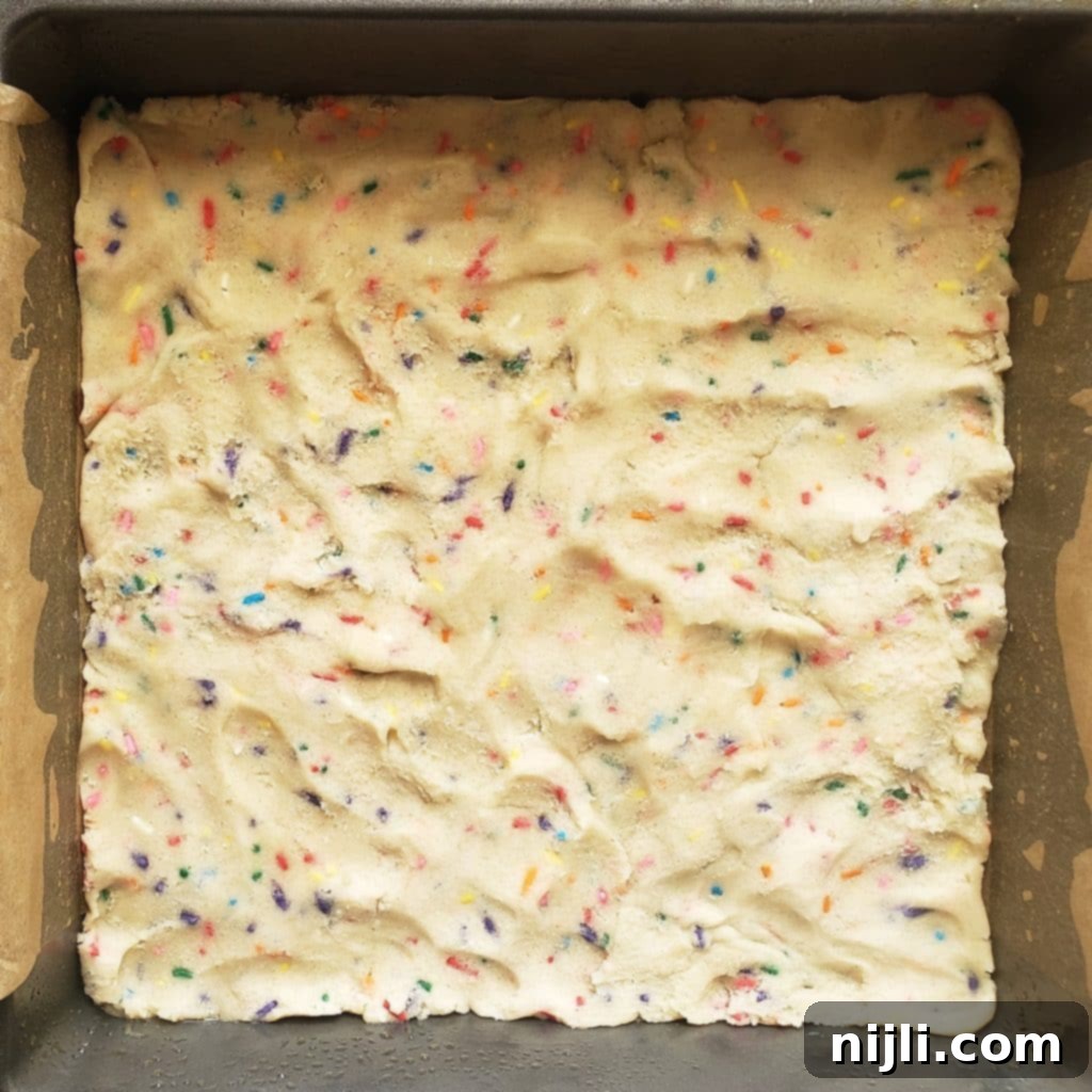 Festive Confetti Cheesecake Bars 4 The initial layer of vibrant funfetti cookie dough meticulously pressed into the bottom of a 9x9 baking pan, forming a sturdy and colorful base for the confetti cheesecake bars.
