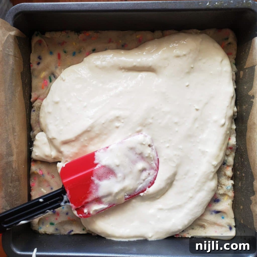 Festive Confetti Cheesecake Bars 5 The smooth, creamy cheesecake filling being carefully spread over the colorful funfetti cookie dough base in a baking pan, creating the second delicious layer.