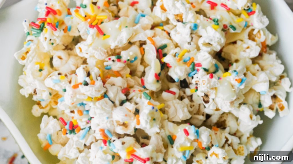 Confetti Popcorn 3 Confetti popcorn spread out on parchment paper, being drizzled with melted white chocolate and topped with rainbow sprinkles.