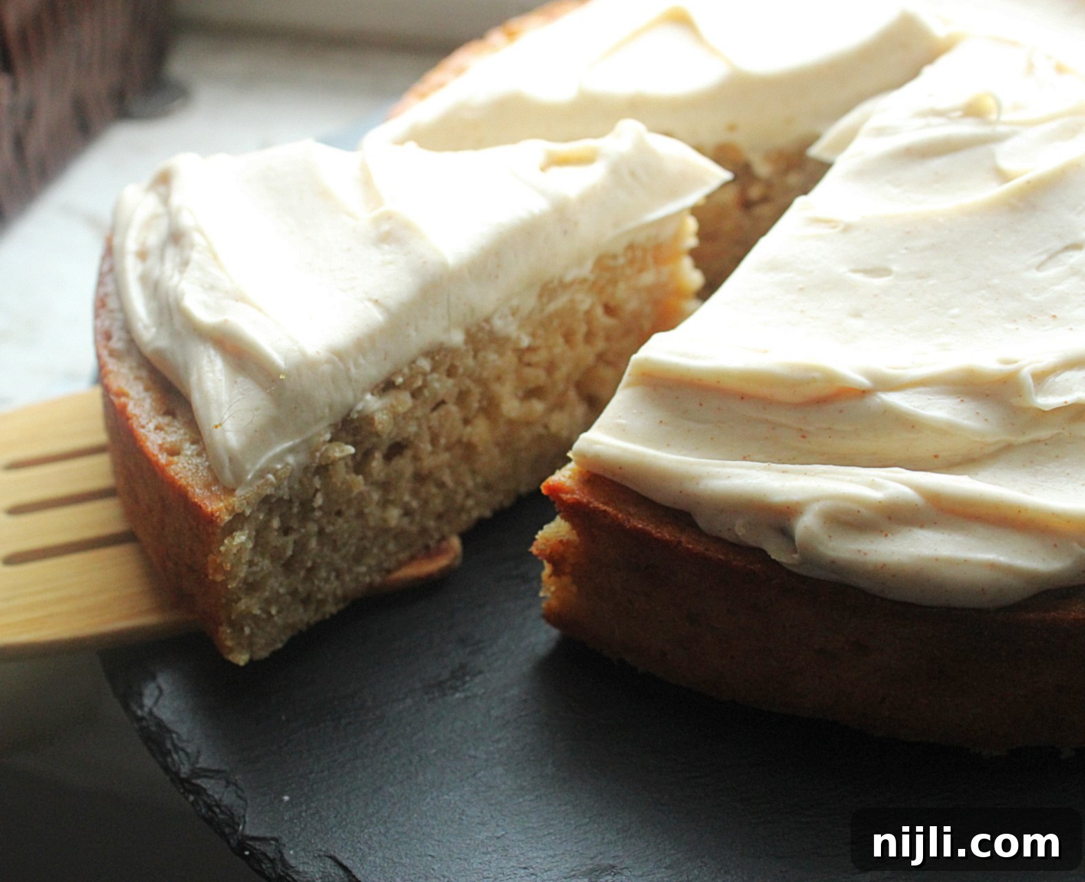 Banana Spice Cake with Toasted Butter Cream Cheese 2 Side angled view of old fashioned spiced banana cake topped with fluffy brown butter cream cheese frosting, with a wooden spatula removing a slice.