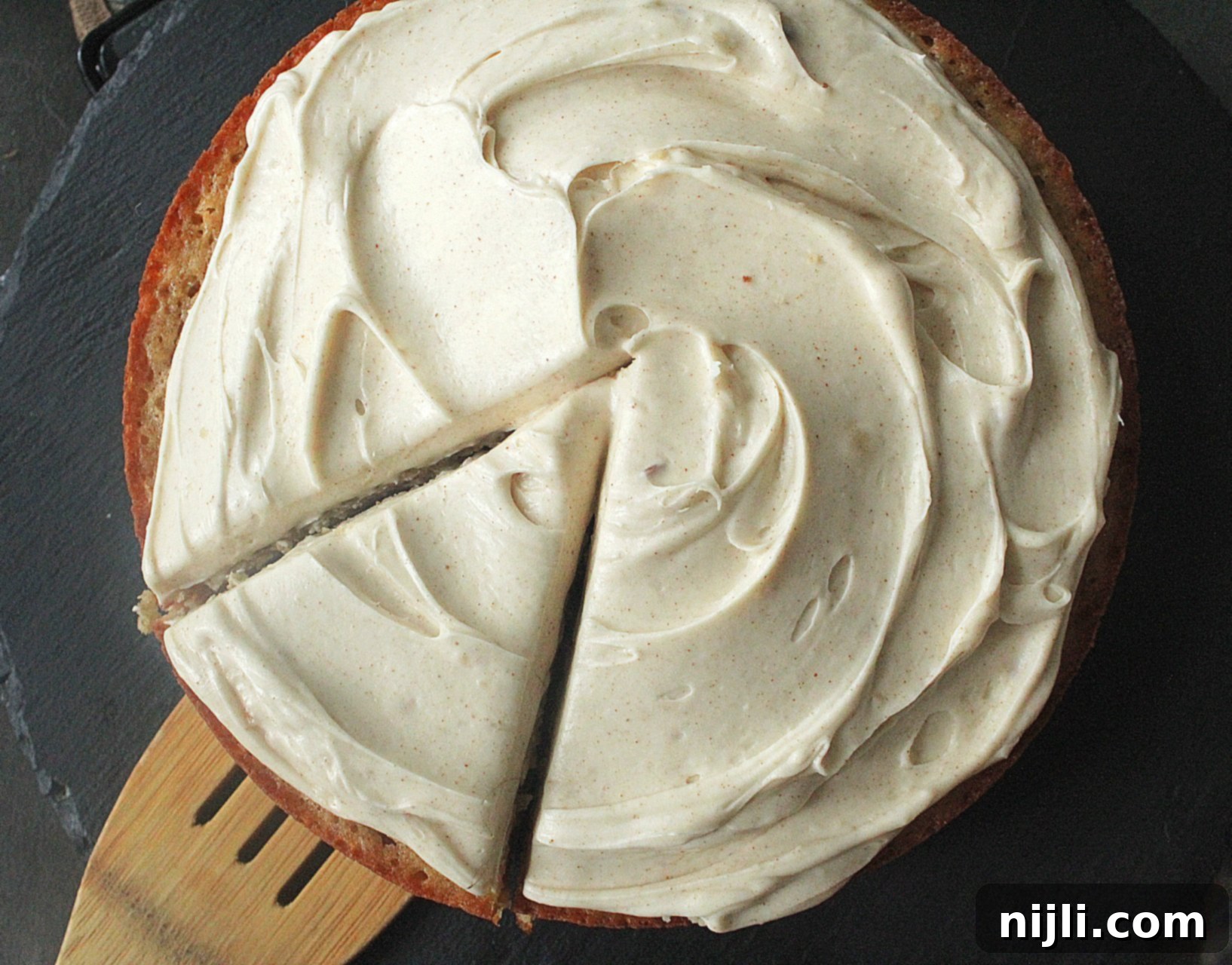 Banana Spice Cake with Toasted Butter Cream Cheese 3 Top view of easy banana spice cake, sitting on a slate serving tray topped with creamy brown butter cream cheese frosting, with a wooden spatula serving a slice.