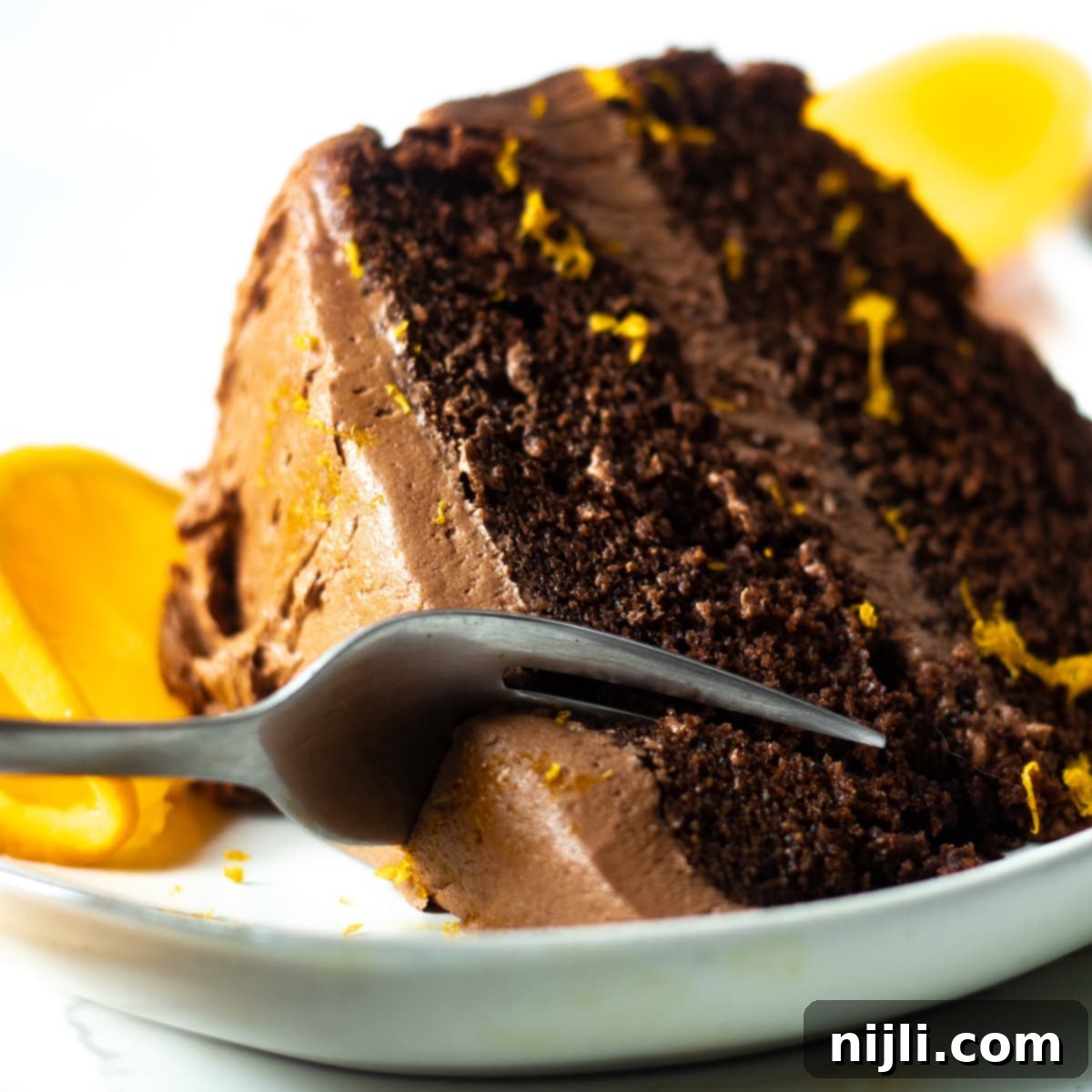 Zesty Chocolate Orange Sensation 7 slice of dark chocolate orange cake being cut with a fork, with an orange and orange zest as garnish