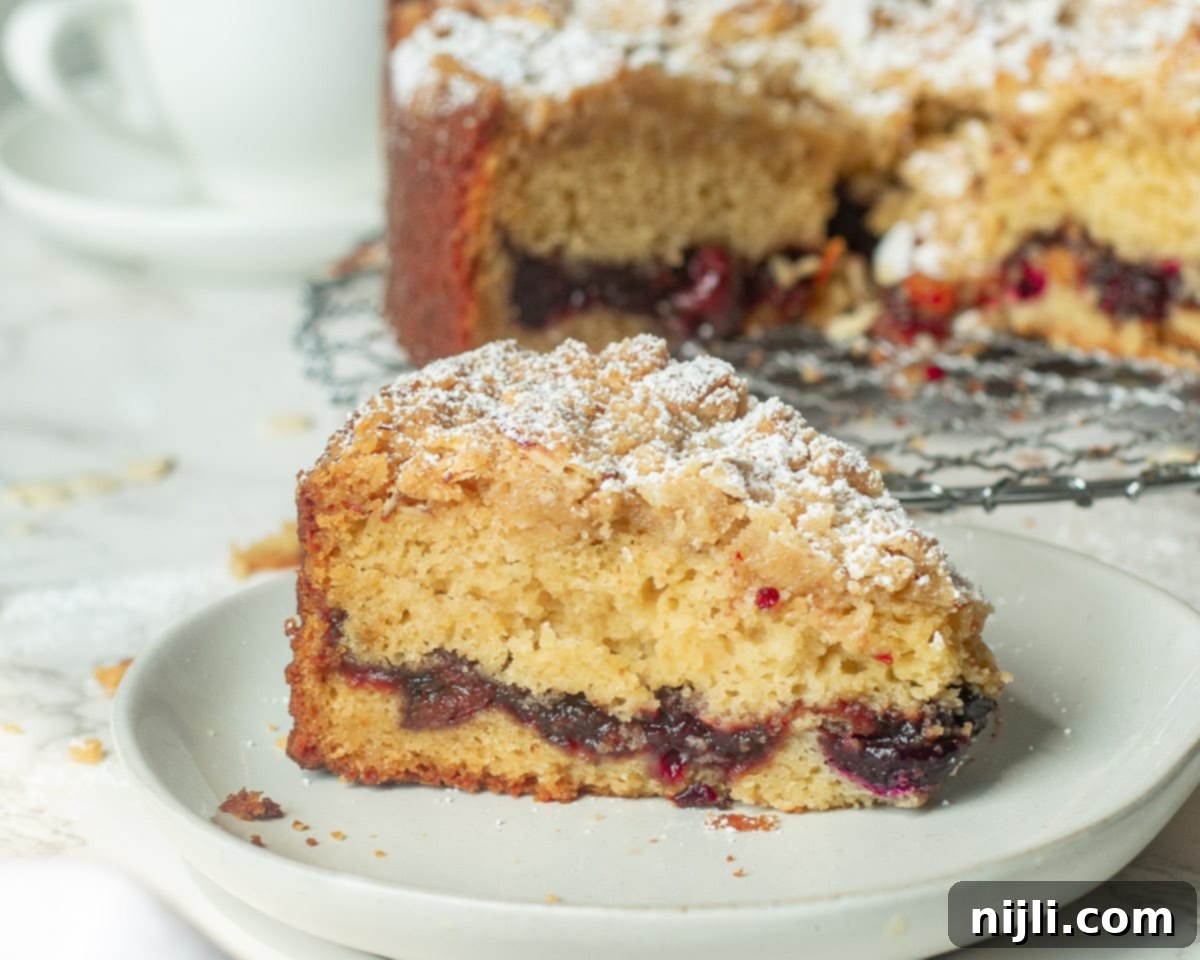 A close-up shot of a freshly cut slice of cherry coffee cake on a plate, revealing the layers of moist cake, rich cherry filling, and crunchy almond crumb topping, dusted with powdered sugar.