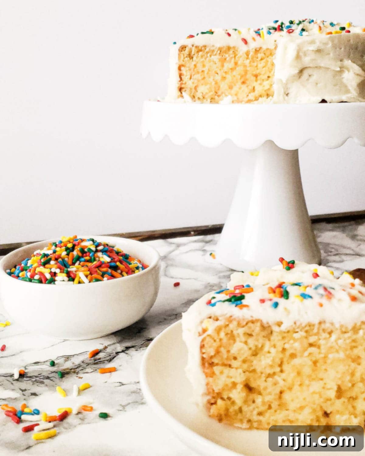 Easy Mini Vanilla Cakes Vanilla Buttercream 5 Mini vanilla cake on a cake stand with a bowl of sprinkles and a slice of cake on a plate, highlighting its delicious simplicity and customization potential.