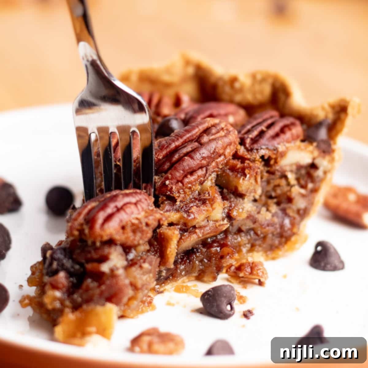Close-up of a slice of chocolate chip pecan pie on a white plate, with a fork cutting through gooey brown sugar filling, melted chocolate chips, and toasted pecans.
