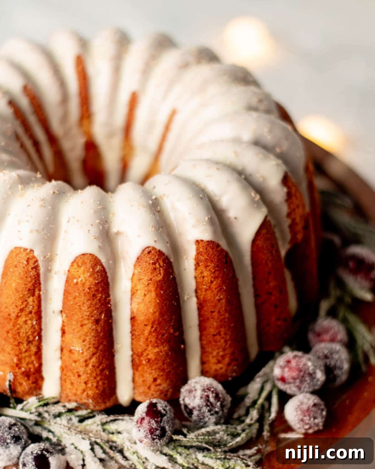 Festive eggnog pound cake covered in white glaze, surrounded by sugared rosemary and cranberries for a Christmas dessert centerpiece.