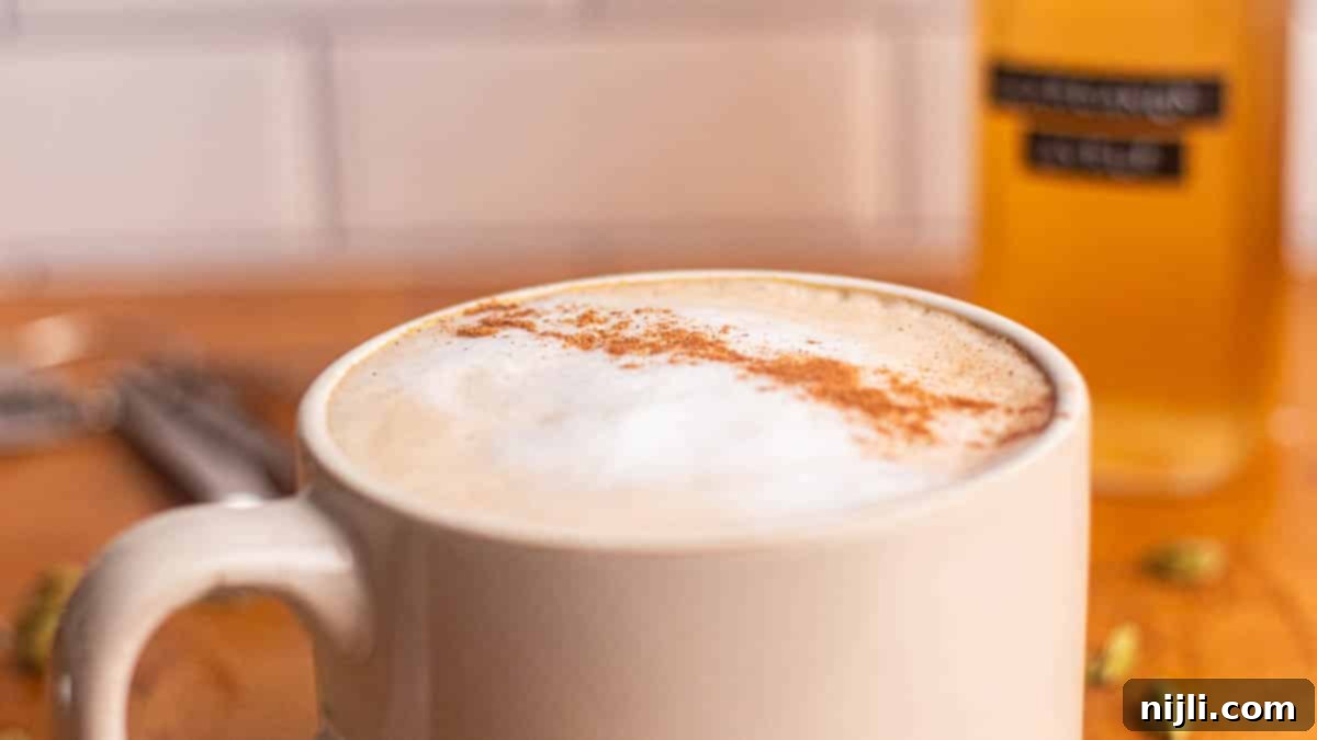 Mug of frothy latte dusted with cinnamon next to a bottle of cardamom simple syrup on a wooden table.