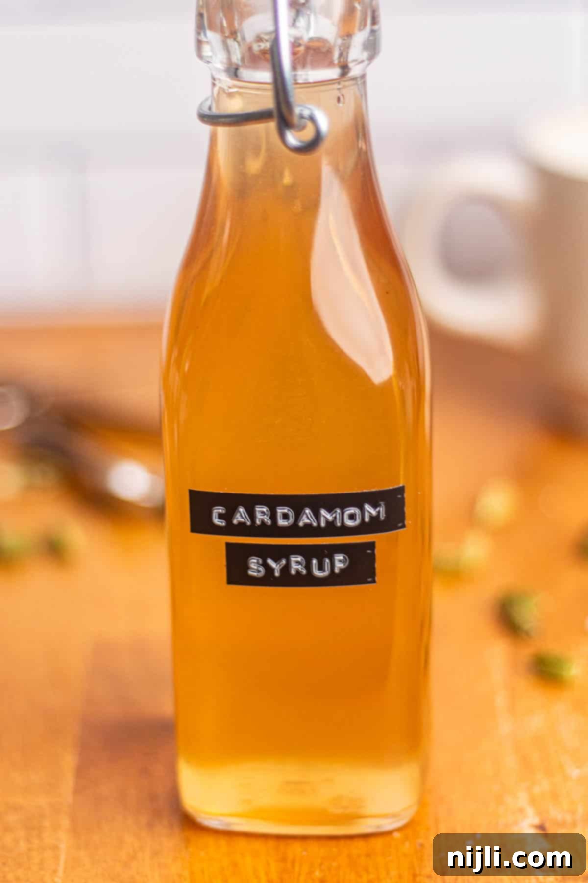 Close-up of a glass bottle of golden brown cardamom syrup on a table with coffee mug in the background.